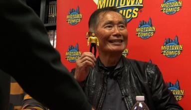 George Takei dressed in a black shirt and black leather jacket sits against a red "Midtown Comics" background while holding up a figurine of his Star Trek character